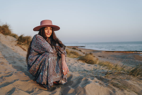 Photograph of model on beach wearing the new Florence maxi dress in powder blue and peach paisley print with balloon sleeves and long flowy skirt, accessorised with light pink felt hat.