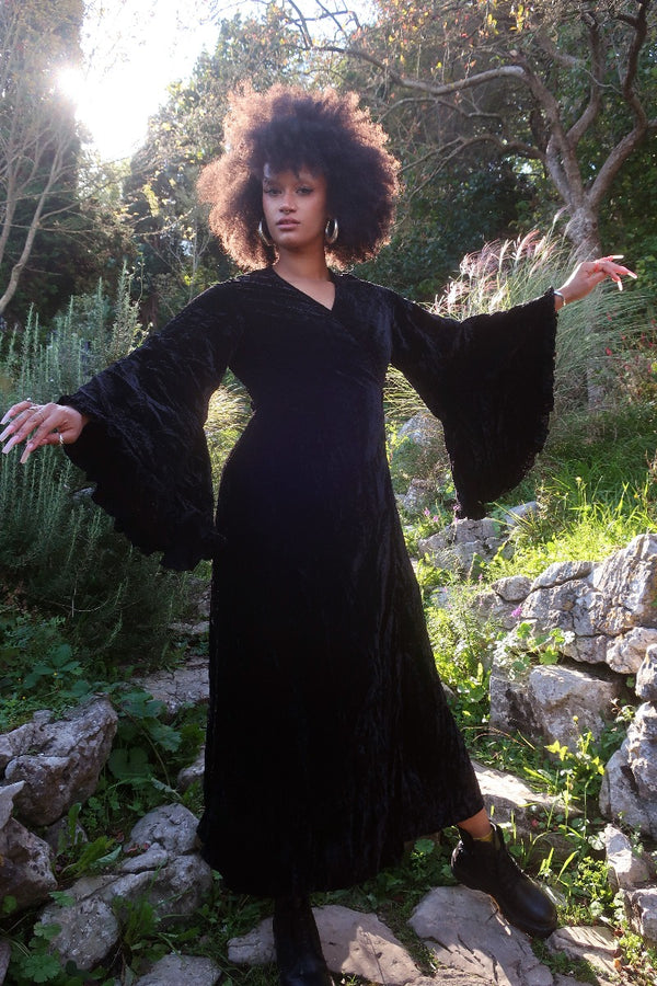 Model wears our Venus Maxi Dress in Morticia Black Velvet. Worn as a wrap style tied at the back with huge floaty bell sleeves. By All About Audrey