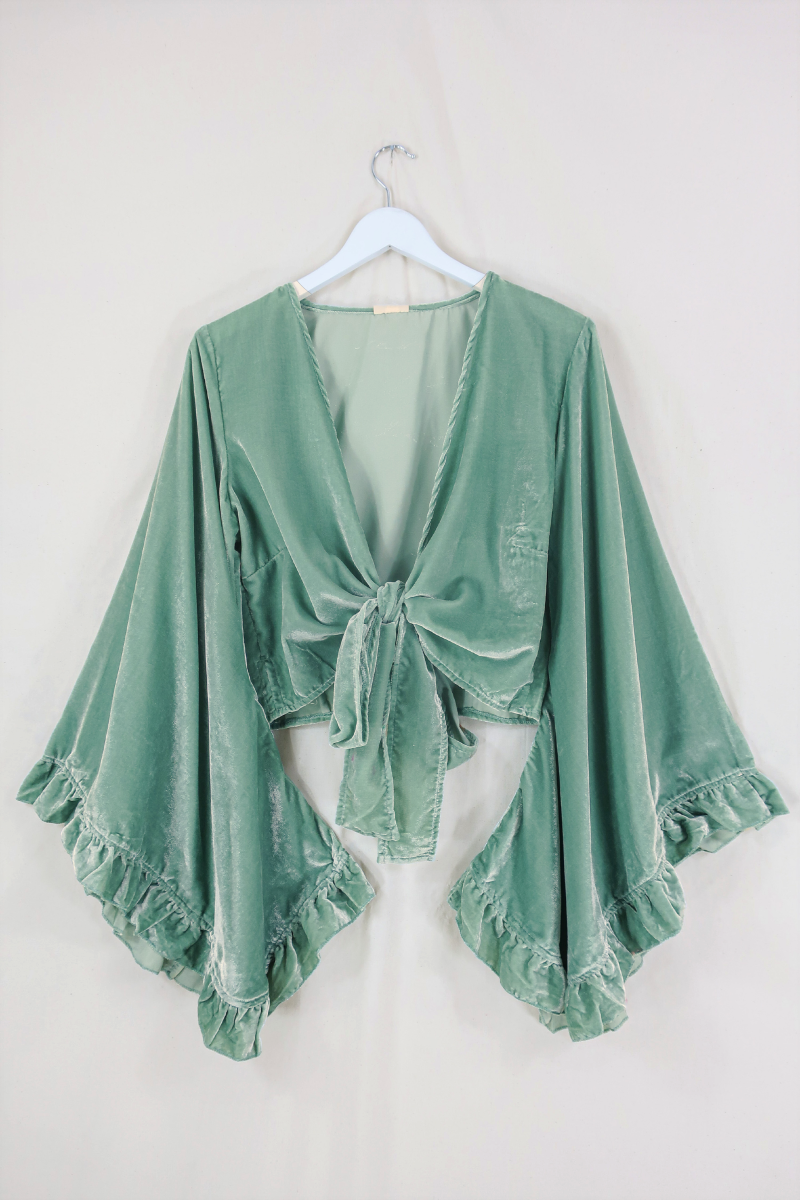 Flat lay of our Velvet Venus Wrap Top in Pistachio Green. A light sage cool tone hue in a soft shimmering velvet. Featuring huge bell sleeves with a frill edge. Showed here tied at the front inspired by 70's bohemia styles. By All About Audrey