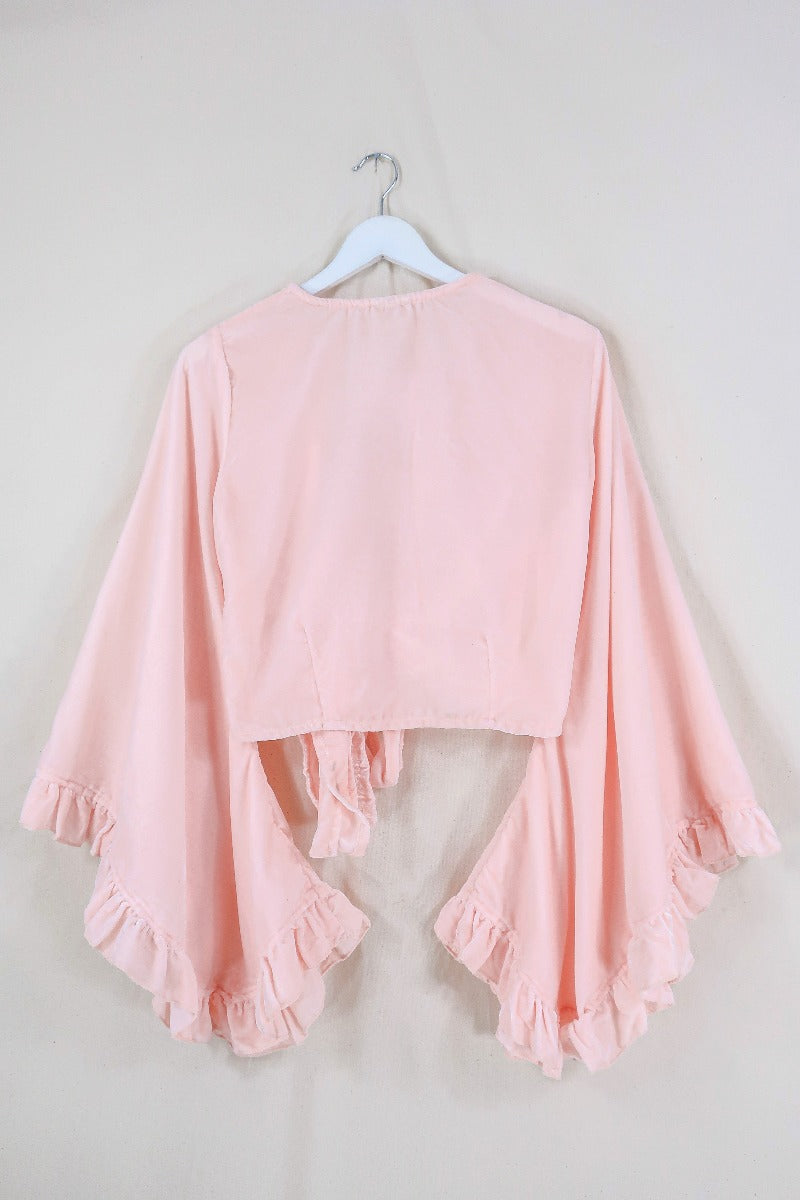 Flat lay of our Velvet Venus Wrap Top in Powder Pink. An ethereal petal pink hue in a soft shimmering velvet. Featuring huge bell sleeves with a frill edge. Shown here tied at the front inspired by 70's bohemia styles. By All About Audrey