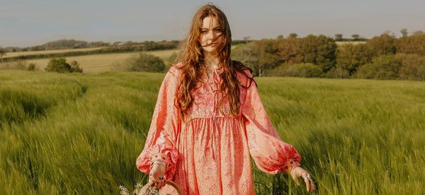 Photograph of model wearing long flowy bohemian dress in pink with big balloon sleeves in a field.