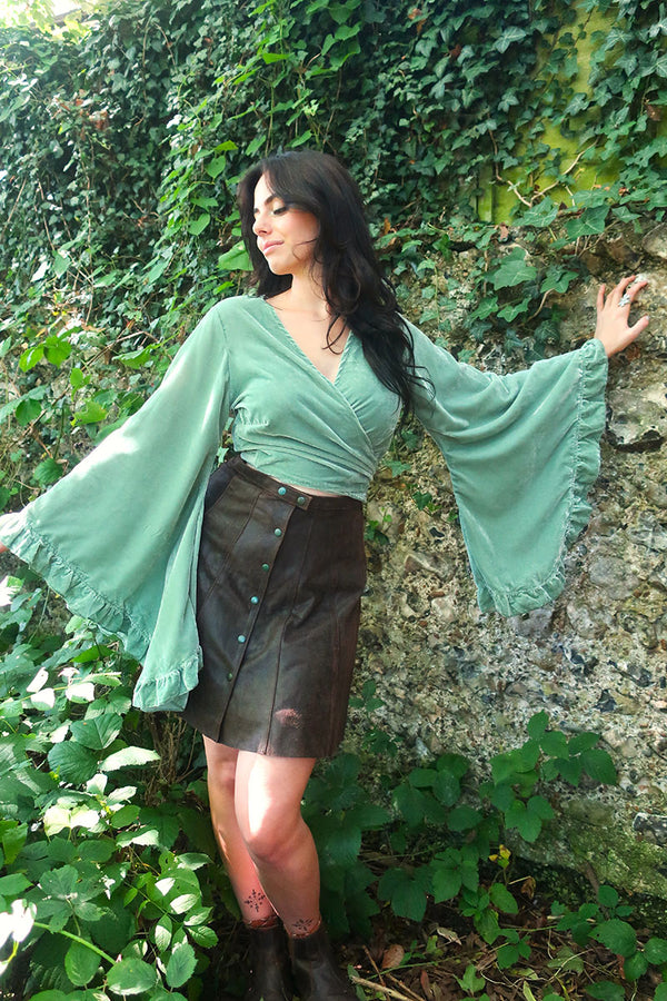 model image of our Velvet Venus Wrap Top in Pistachio Green. A light sage cool tone hue in a soft shimmering velvet. Featuring huge bell sleeves with a frill edge. Showed here tied at the front inspired by 70's bohemia styles. By All About Audrey