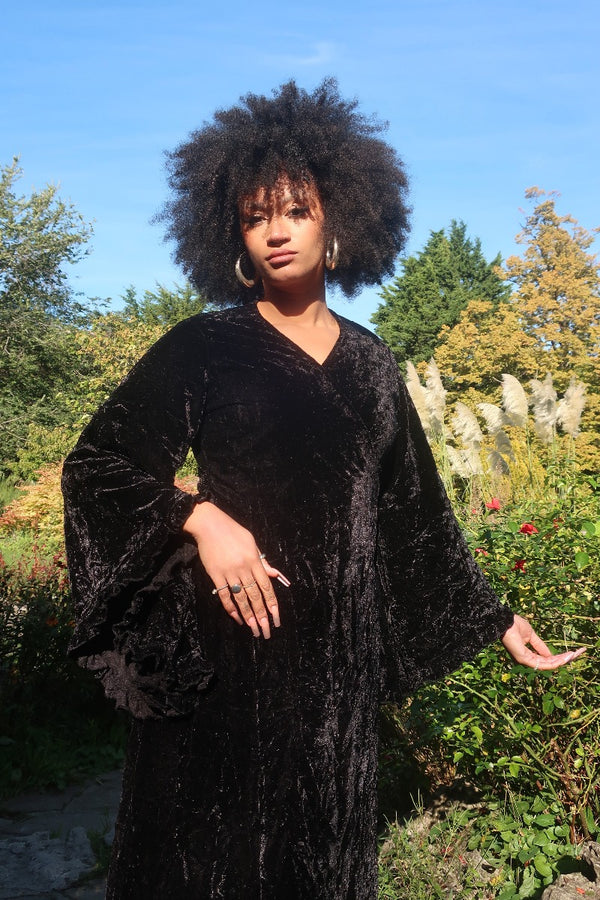 Model wears our Venus Maxi Dress in Morticia Black Velvet. Worn as a wrap style tied at the back with huge floaty bell sleeves. By All About Audrey