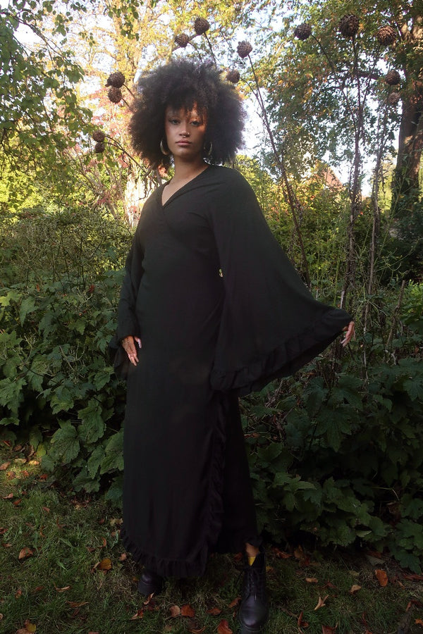 Model wears our Khroma Venus Robe Dress in Nightshade Black in a wrap dress style. A 70's boho inspired design with frills on the sleeves and the front. By All About Audrey