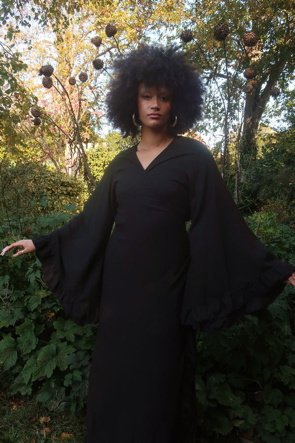 Model wears our Khroma Venus Robe Dress in Nightshade Black in a wrap dress style. A 70's boho inspired design with frills on the sleeves and the front. By All About Audrey