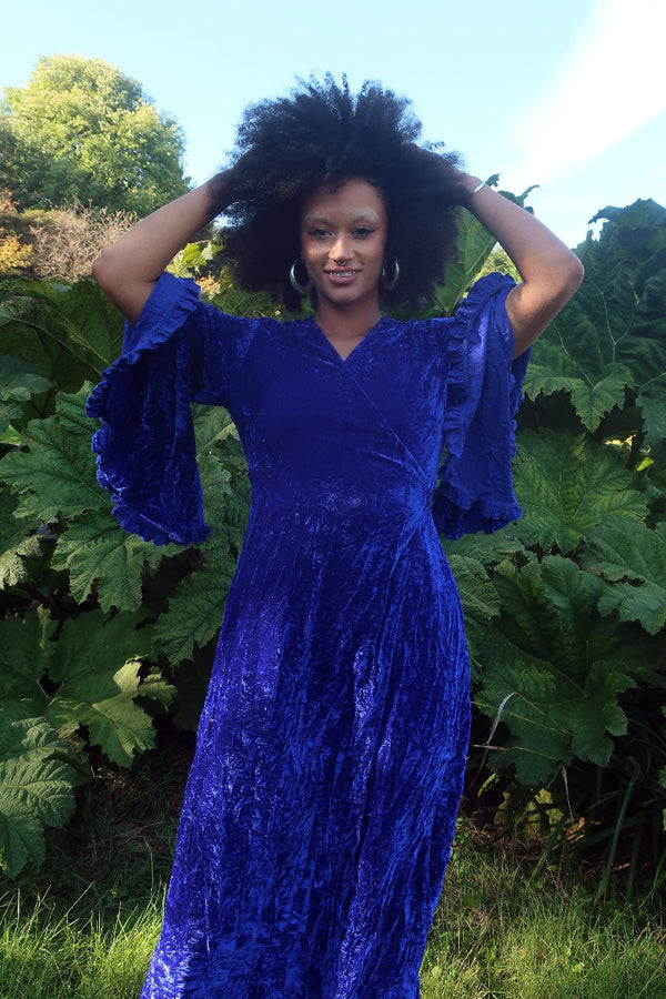 Model wears our Khroma Venus Maxi Dress in Regal Blue Velvet. Showing off the vintage 1970s inspired bell sleeves and worn in a wrap style, tied at the back. By All About Audrey