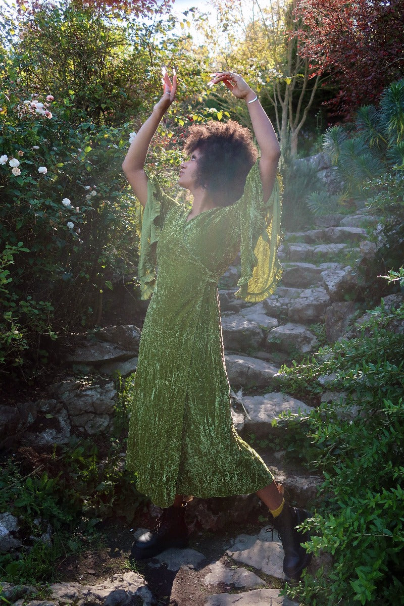 Model wears our Khroma Venus Maxi Dress in Elven Green Velvet. Worn like a wrap style and tied at the back with huge floaty retro inspired bell sleeves. By All About Audrey