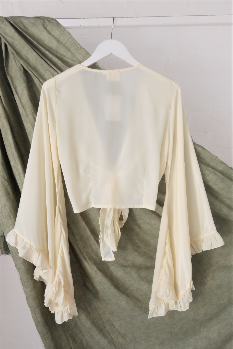 A flat lay photo of a floaty and ethereal bell sleeve wrap top in a cream off white colourway inspired by 1970s bohemia fashion by All About Audrey