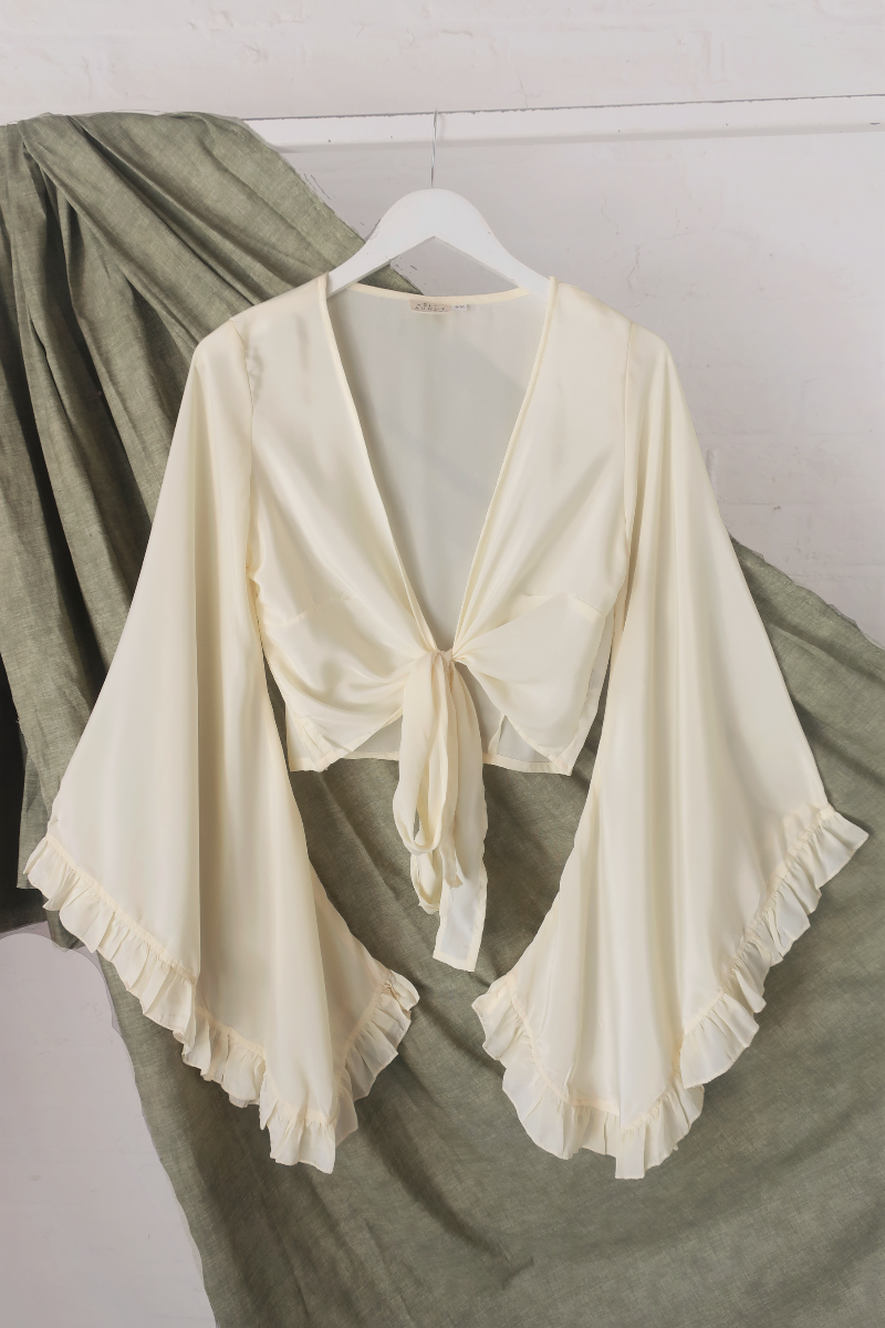 A flat lay photo of a floaty and ethereal bell sleeve wrap top in a cream off white colourway inspired by 1970s bohemia fashion by All About Audrey