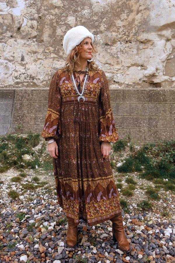 Audrey wears our Peacock Primrose Dress in Gingerbread. A dark brown tone with an earth tone peacock and floral print. Inspired by 1970s bohemia, paired with chunky boho jewellery and our Anastasia hat. All by All About Audrey