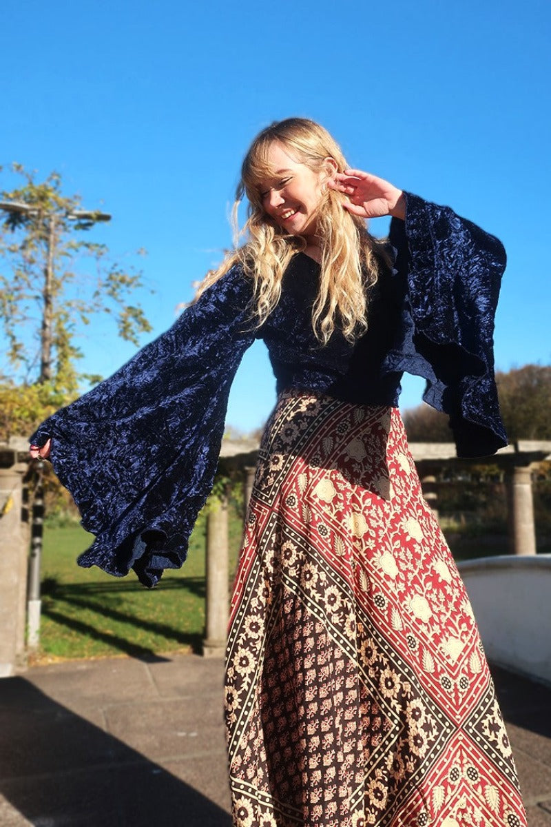 model styling one of our Velvet Venus Wrap Top in galaxy blue. A vast dark starry blue tone in a soft shimmering velvet. Featuring huge bell sleeves with a frill edge. Shown tied at the front inspired by 70's bohemia styles. By All About Audrey