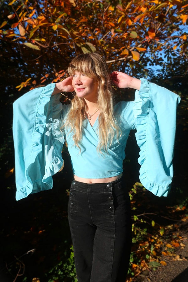 model styling one of our Velvet Venus Wrap Top in Sky Blue. A retro bold aqua blue hue in a soft shimmering velvet. Featuring huge bell sleeves with a frill edge. Shown tied at the front inspired by 70's bohemia styles. By All About Audrey