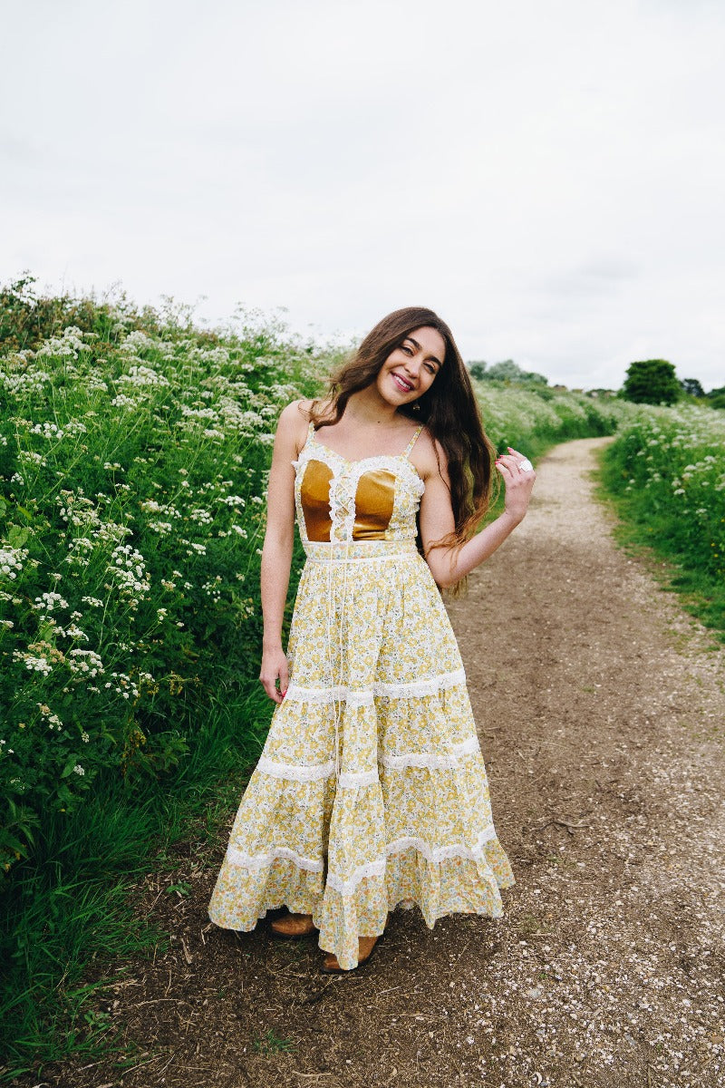 Full length model photo of Darcy dress in Saffron Bloom. With a repeated bohemian romantic ditsy yellow floral print throughout and prairie lace detailing. Elegant fitted bodice with corset fastening and full frill tiered maxi skirt By All About Audrey