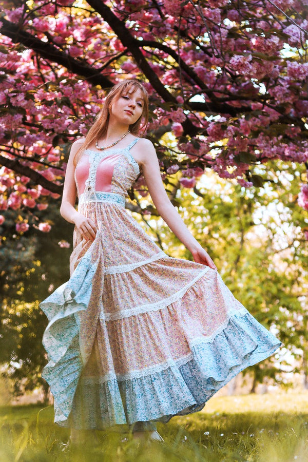 Full length model photo of Darcy dress in Wild Strawberry Pink. With a repeated bohemian romantic ditsy yellow floral print throughout and prairie lace detailing. Elegant fitted bodice with corset fastening and full frill tiered maxi skirt By All About Audrey