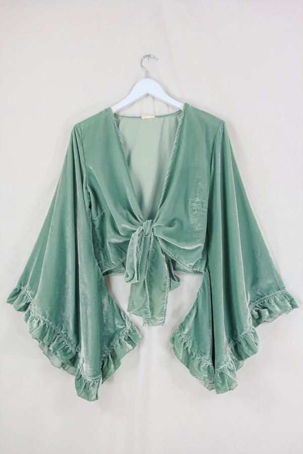 Flat lay of our Velvet Venus Wrap Top in Pistachio Green. A light sage cool tone hue in a soft shimmering velvet. Featuring huge bell sleeves with a frill edge. Showed here tied at the front inspired by 70's bohemia styles. By All About Audrey