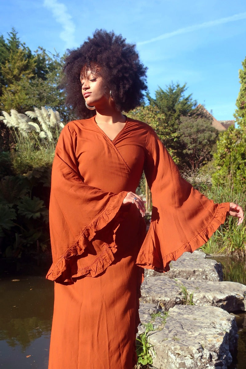 Model wears our Khroma Venus Robe Dress in Red Clay as a wrap style dress, showing off the huge bell sleeves and frill trim on the sleeves and front. All About Audrey