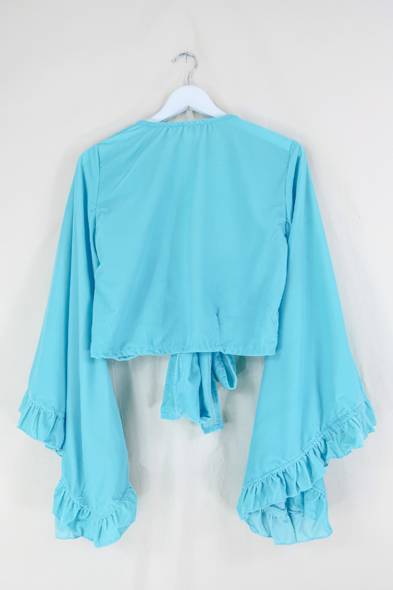 Flat lay of our Velvet Venus Wrap Top in Sky Blue. A retro bold aqua blue hue in a soft shimmering velvet. Featuring huge bell sleeves with a frill edge. Shown tied at the front inspired by 70's bohemia styles. By All About Audrey