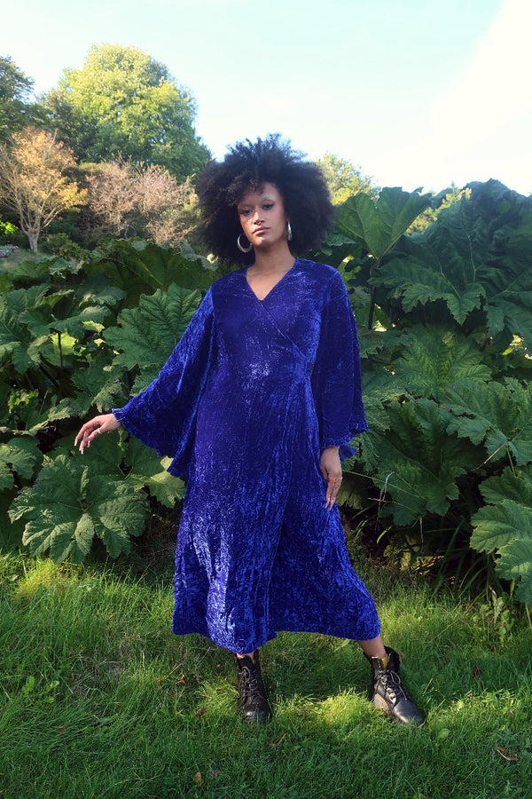 Model wears our Khroma Venus Maxi Dress in Regal Blue Velvet. Showing off the vintage 1970s inspired bell sleeves and worn in a wrap style, tied at the back. By All About Audrey
