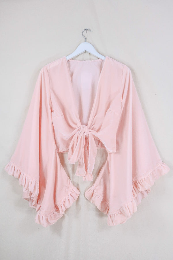 Flat lay of our Velvet Venus Wrap Top in Powder Pink. An ethereal petal pink hue in a soft shimmering velvet. Featuring huge bell sleeves with a frill edge. Shown here tied at the front inspired by 70's bohemia styles. By All About Audrey