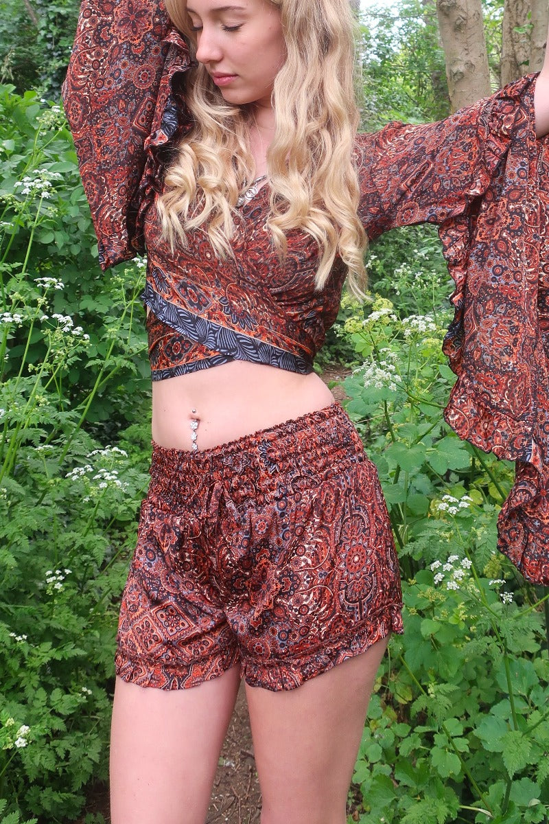 Photo shows model wearing our Venus frilly shorts in rust and jet mandala. The print is a mandala tile print with black, rust with stone blue details. The waist band is ruched with a tie belt. These shorts are free size.