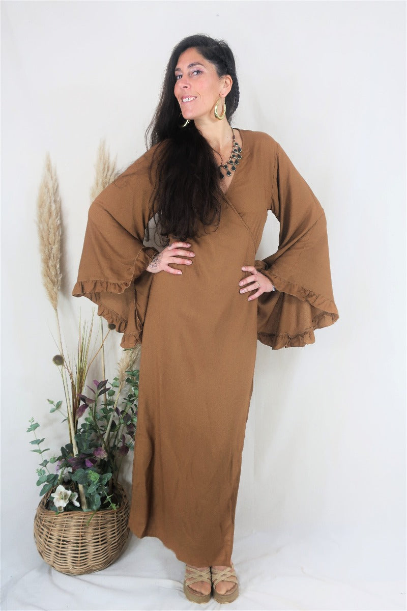 Model wears our bohemian bell sleeve wrap maxi dress in cinnamon brown. Long floaty sleeves with a ruffle hem and a wraparound tie waist to create an elegant shape by all about audrey