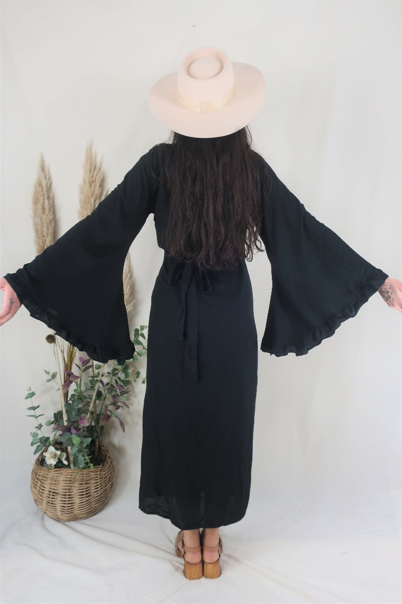 Back full photo of the model wearing our Venus rayon 70s inspired maxi bell sleeve wrap dress in Nightshade Black. Model shows off the oversized butterfly sleeves and adjustable wrap tie waist by All About Audrey